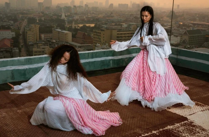 Two women in flowing striped dresses pose on a rooftop overlooking a cityscape at sunset, creating a fashion editorial-style image.
