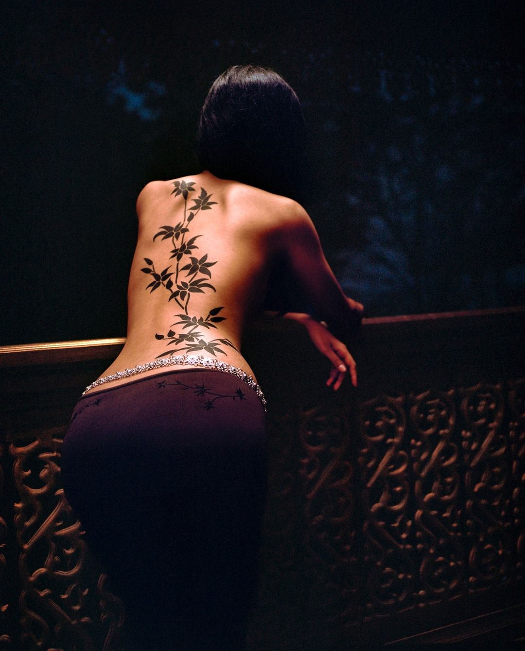 Adult female stands at ornate railing with a back tattoo featuring botanical illustration of leaves and branches reaching up her back.
