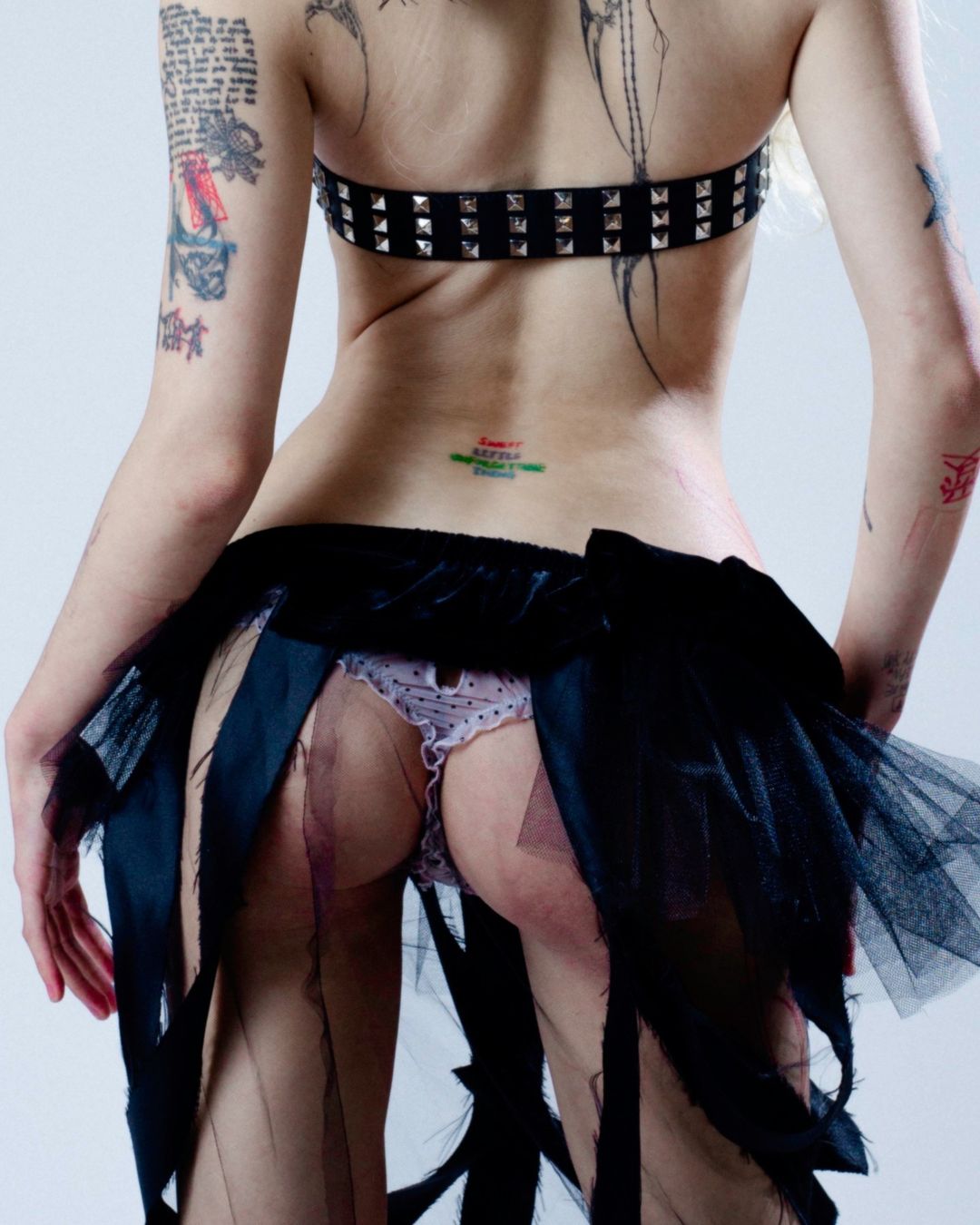 Back view of an adult woman with tattoos, wearing a studded bra and a black tulle skirt, posing in a bright photo studio.