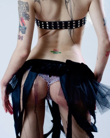 Rear view of a person with numerous tattoos wearing a studded belt, black ruffled skirt, and patterned underwear against a white background