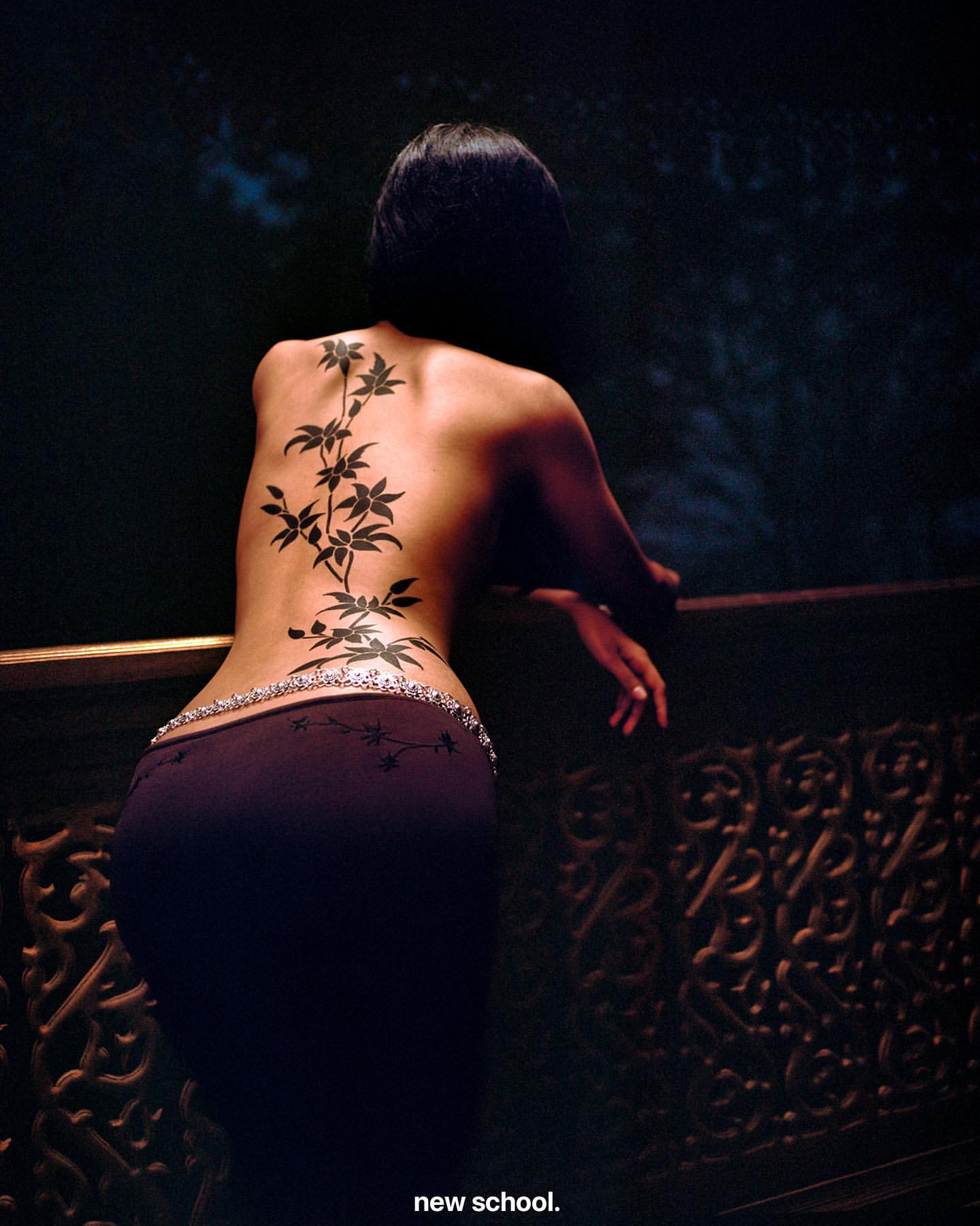 Back view of a woman with a large botanical tattoo design on her back, wearing low-rise pants with beaded detailing on the waistband.