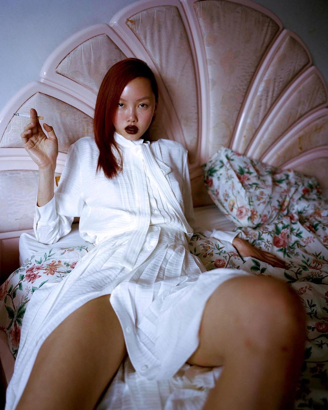 Asian woman with auburn hair in a white shirt and skirt sitting on a floral bed while holding a cigarette in an indoor setting.