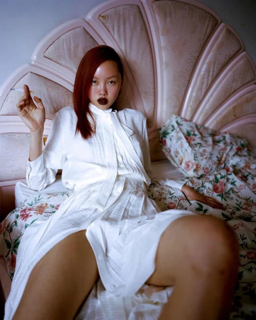 Asian woman with auburn hair in a white shirt and skirt sitting on a floral bed while holding a cigarette in an indoor setting.