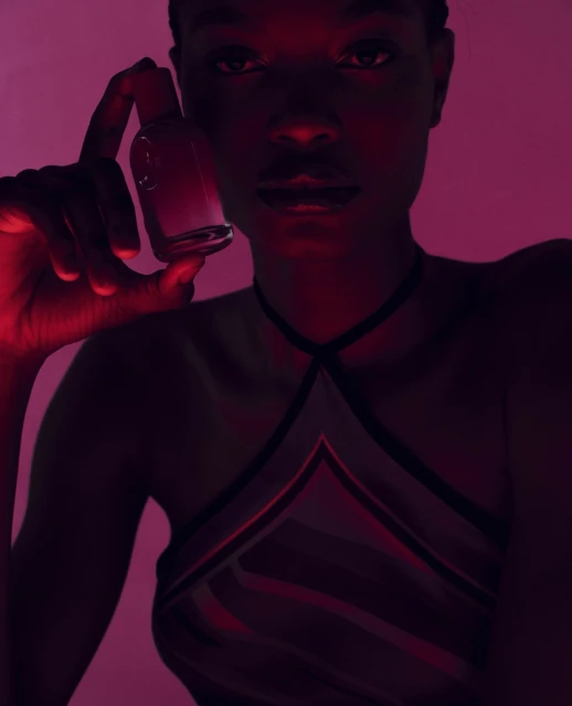 Close-up studio shot of a young woman holding a translucent perfume bottle, bathed in dramatic red and pink light against a plain backdrop.