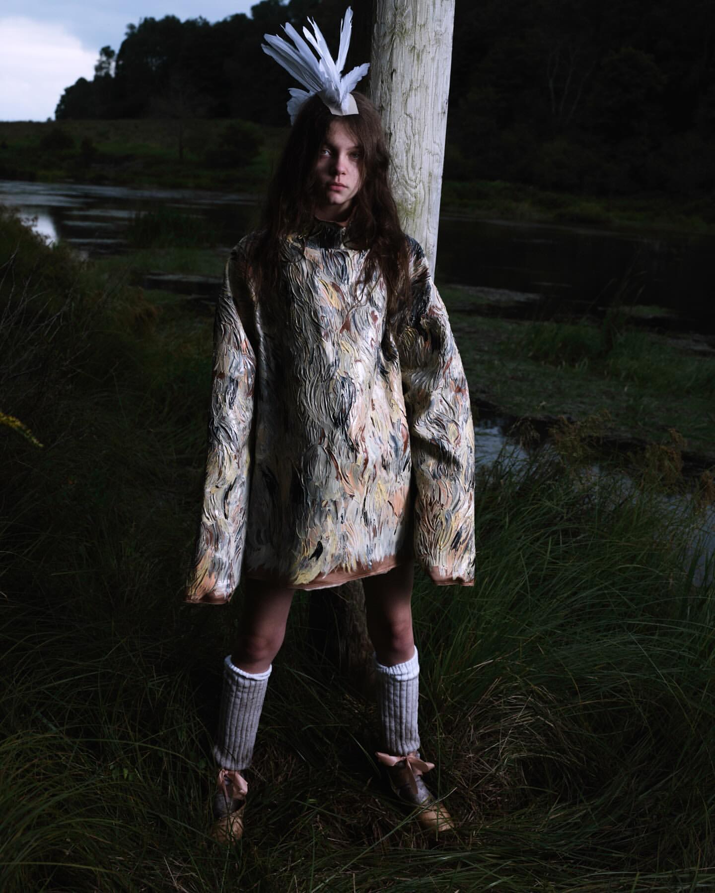 A young individual in an artistic outfit with a feathered headpiece poses outdoors in a grassy area near a body of water, creating an editorial fashion image.