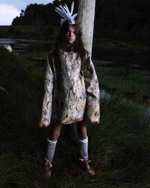 A young individual in an artistic outfit with a feathered headpiece poses outdoors in a grassy area near a body of water, creating an editorial fashion image.