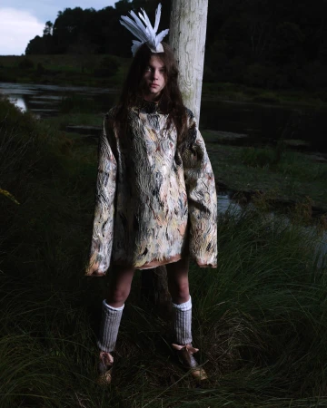 A young individual in an artistic outfit with a feathered headpiece poses outdoors in a grassy area near a body of water, creating an editorial fashion image.