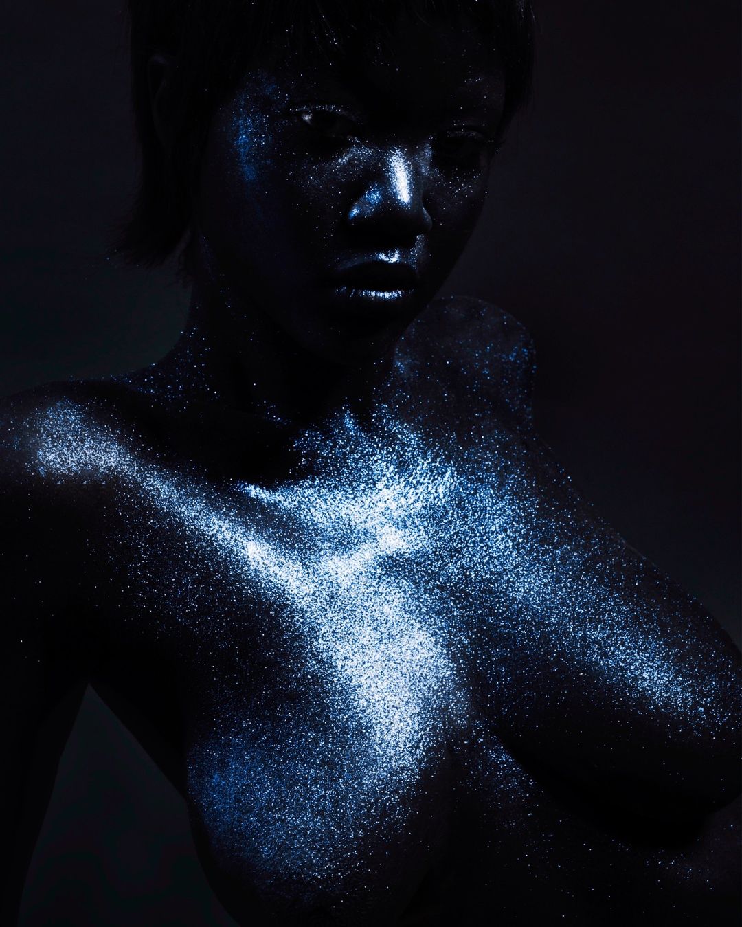 Glamorous dark-skinned woman with shimmering blue makeup and hair, posing against a dark background in a portrait photograph.