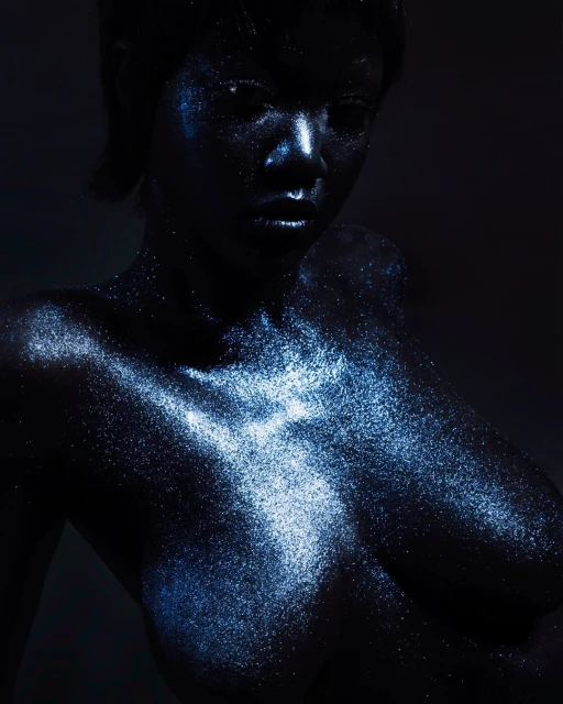 Glamorous dark-skinned woman with shimmering blue makeup and hair, posing against a dark background in a portrait photograph.