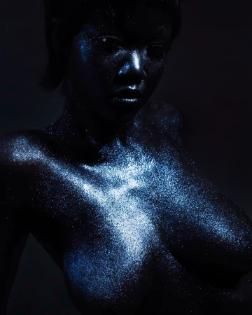 Glamorous dark-skinned woman with shimmering blue makeup and hair, posing against a dark background in a portrait photograph.