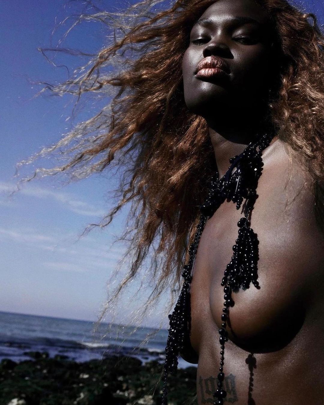 Outdoor portrait of a Black woman with wind-blown curly hair, wearing a beaded necklace, posing against a sky and ocean backdrop.