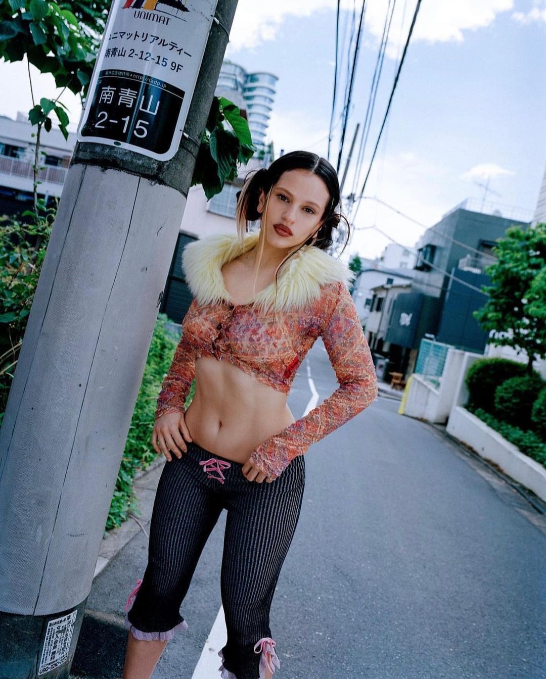 A fashionable young woman poses on a city street in a sheer top with a fur collar and pinstripe pants, standing next to a utility pole.