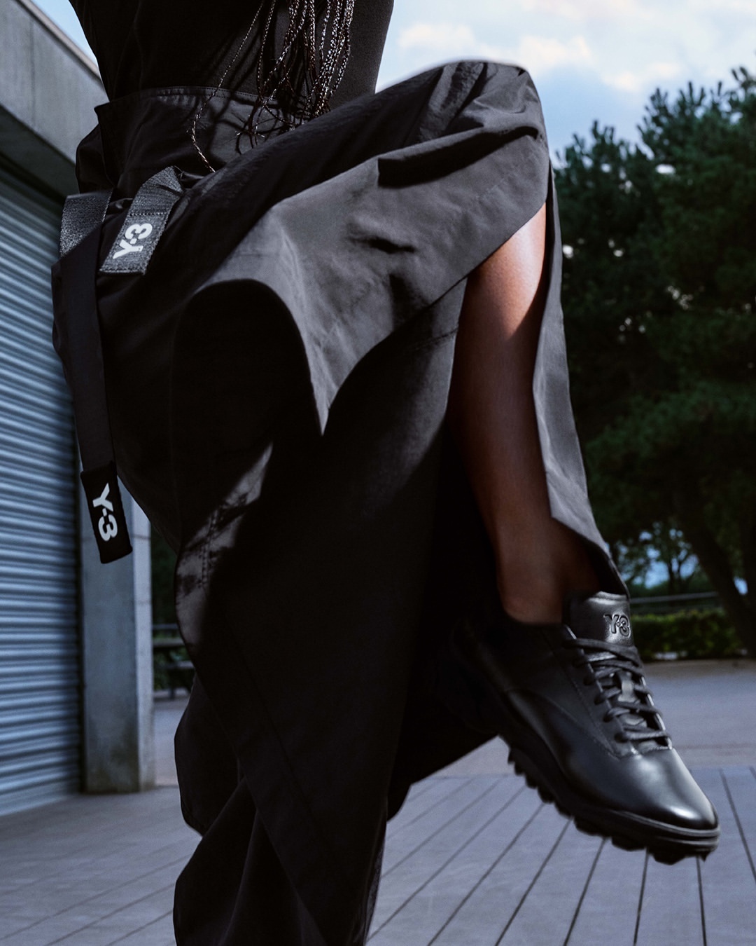 Fashion shot featuring a woman's leg and black sneaker, set against a blurred outdoor backdrop. Y-3 logo is visible on a black belt.
