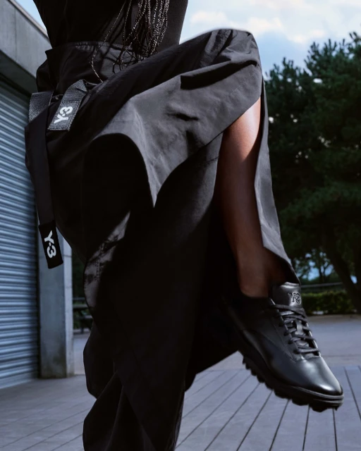 Fashion shot featuring a woman's leg and black sneaker, set against a blurred outdoor backdrop. Y-3 logo is visible on a black belt.