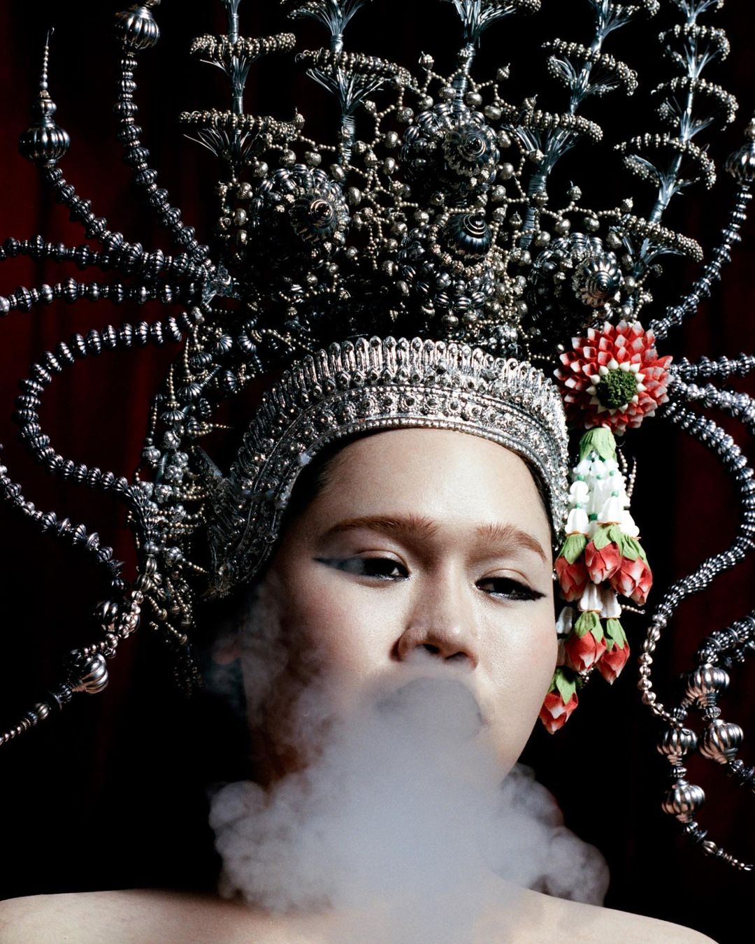 A person with an elaborate metallic headpiece exhales a cloud of smoke, set against a dark red backdrop, creating a dramatic portrait.