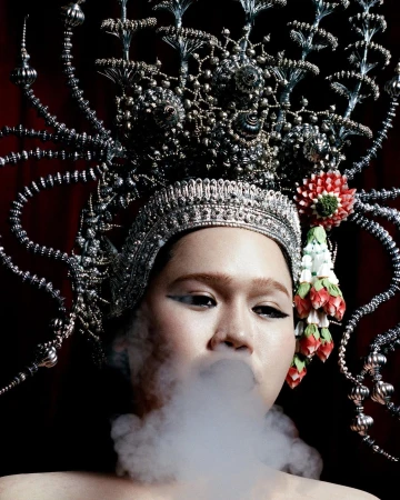 A person with an elaborate metallic headpiece exhales a cloud of smoke, set against a dark red backdrop, creating a dramatic portrait.