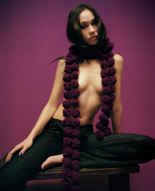 Portrait of a young woman with dark hair wearing jeans and a maroon pompom scarf sitting on a wood stool against a purple background.