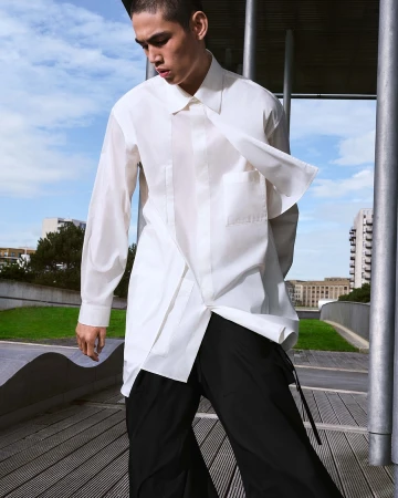 A man in a white button-down shirt and black trousers standing outdoors near metallic pillars under a blue sky.
