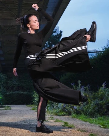 Woman in black athletic top and long skirt kicking in midair outdoors under a concrete bridge by Erika Kamano