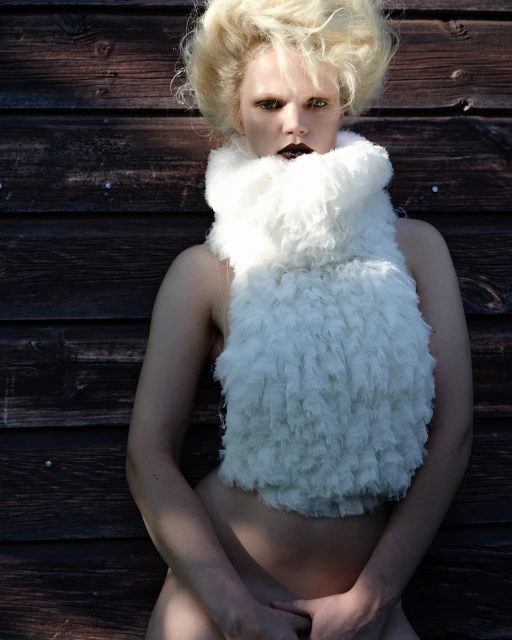 Artistic portrait of a person with voluminous blonde hair and a white, fluffy scarf, posing against a dark wooden background in natural light.