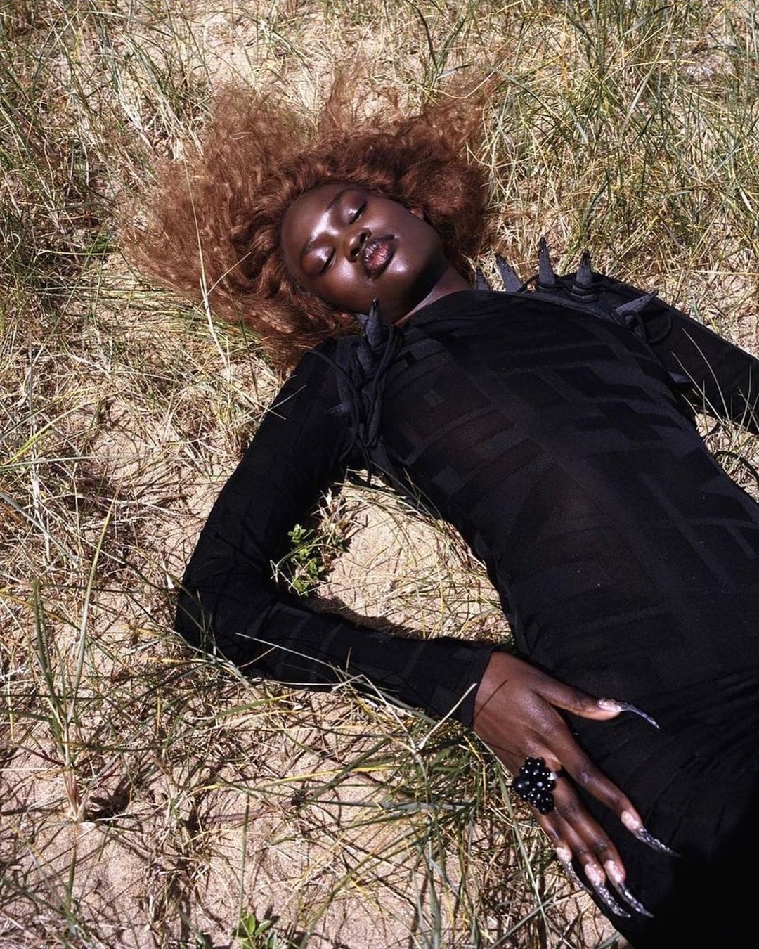 Woman in black dress and ring lying in grassy field with tan, windswept hair in outdoor fashion or editorial photography.