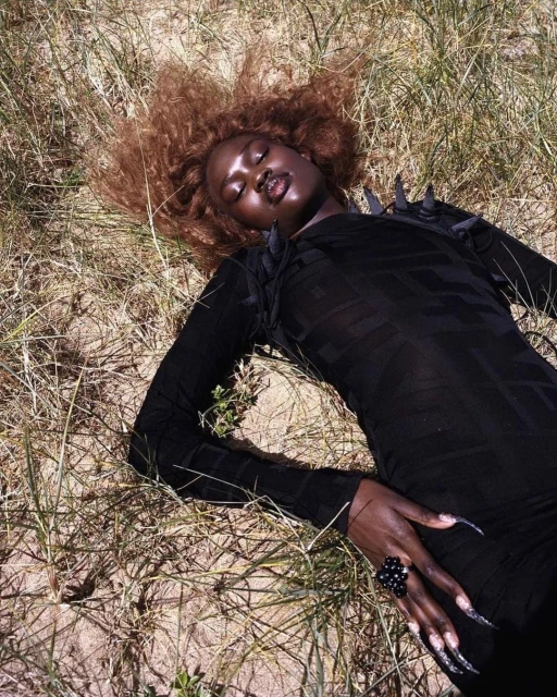 Woman in black dress and ring lying in grassy field with tan, windswept hair in outdoor fashion or editorial photography.