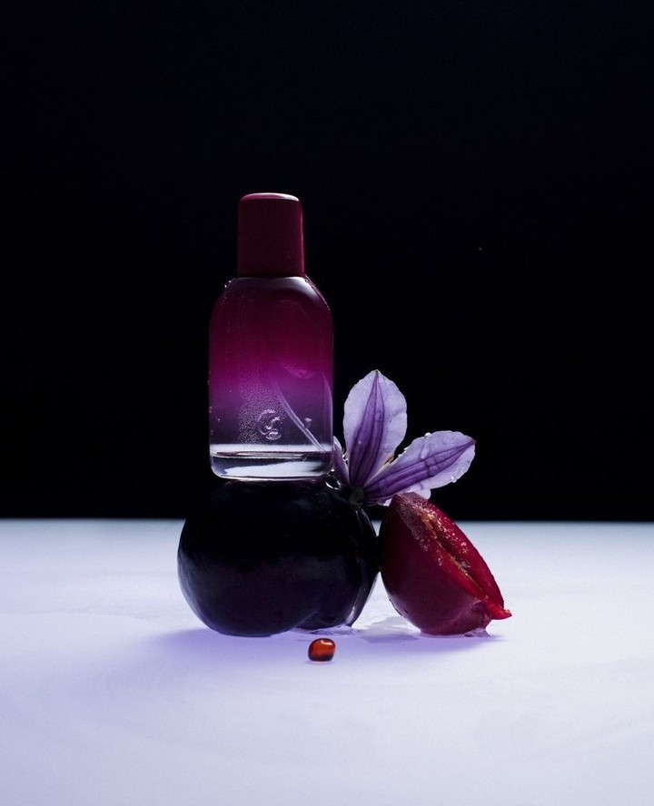 Perfume bottle with violet and plum, studio shot. Gradient color bottle displayed with plum and flower on white glossy background.