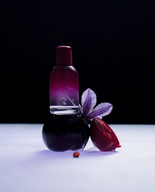 Perfume bottle with violet and plum, studio shot. Gradient color bottle displayed with plum and flower on white glossy background.