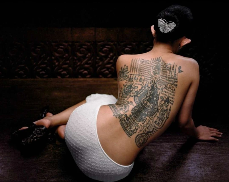Back view of a woman with a large back tattoo wearing white bottoms and black shoes sitting against a dark backdrop with an ornate design.