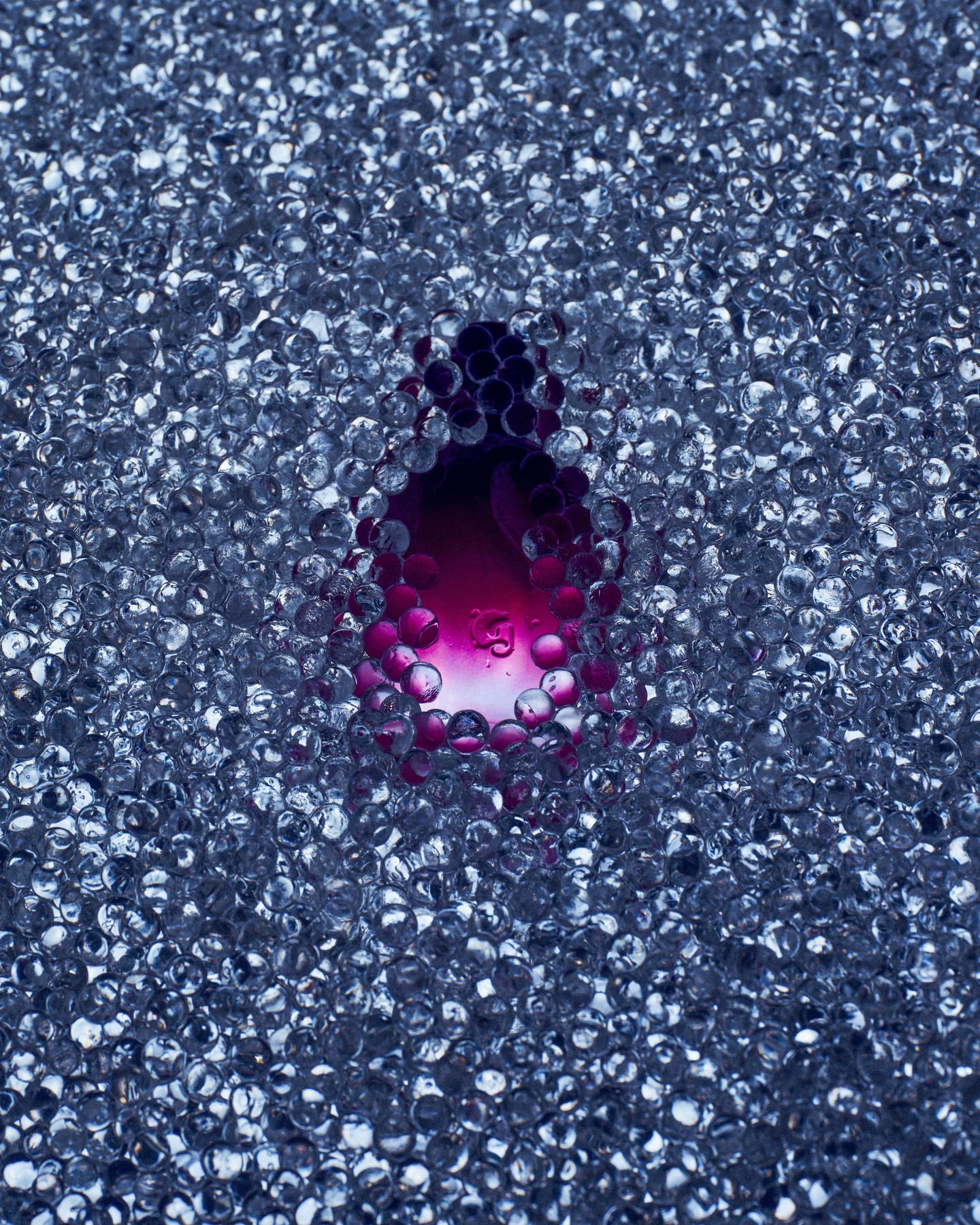 A clear orb surrounds a pool of magenta liquid, resting upon a bed of many small, clear, iridescent, spherical beads with diffused lighting.