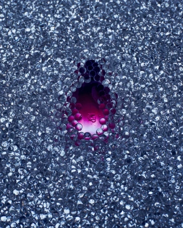 A clear orb surrounds a pool of magenta liquid, resting upon a bed of many small, clear, iridescent, spherical beads with diffused lighting.