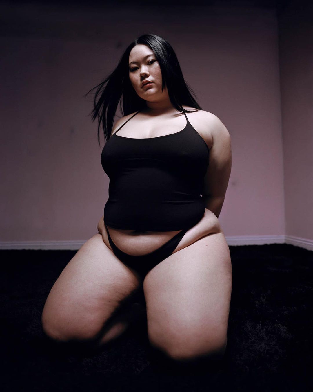 Woman with long dark hair poses kneeling on black shag carpet in a photo studio with pink-tinged walls in a portrait style photo.