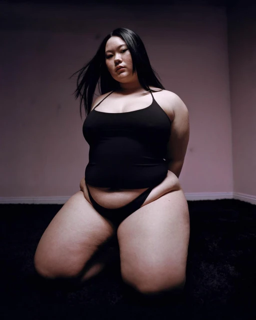 Woman with long dark hair poses kneeling on black shag carpet in a photo studio with pink-tinged walls in a portrait style photo.
