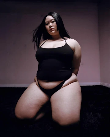 Woman with long dark hair poses kneeling on black shag carpet in a photo studio with pink-tinged walls in a portrait style photo.