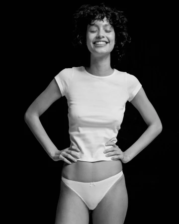 Monochrome portrait of a smiling woman with short curly hair wearing a white t-shirt and white underwear against a black background.