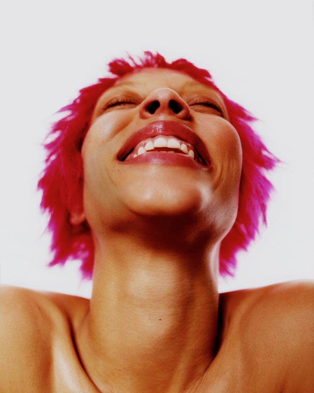 Close-up low-angle portrait of a woman with vibrant pink hair and glossy lips smiling with eyes closed against a white background.