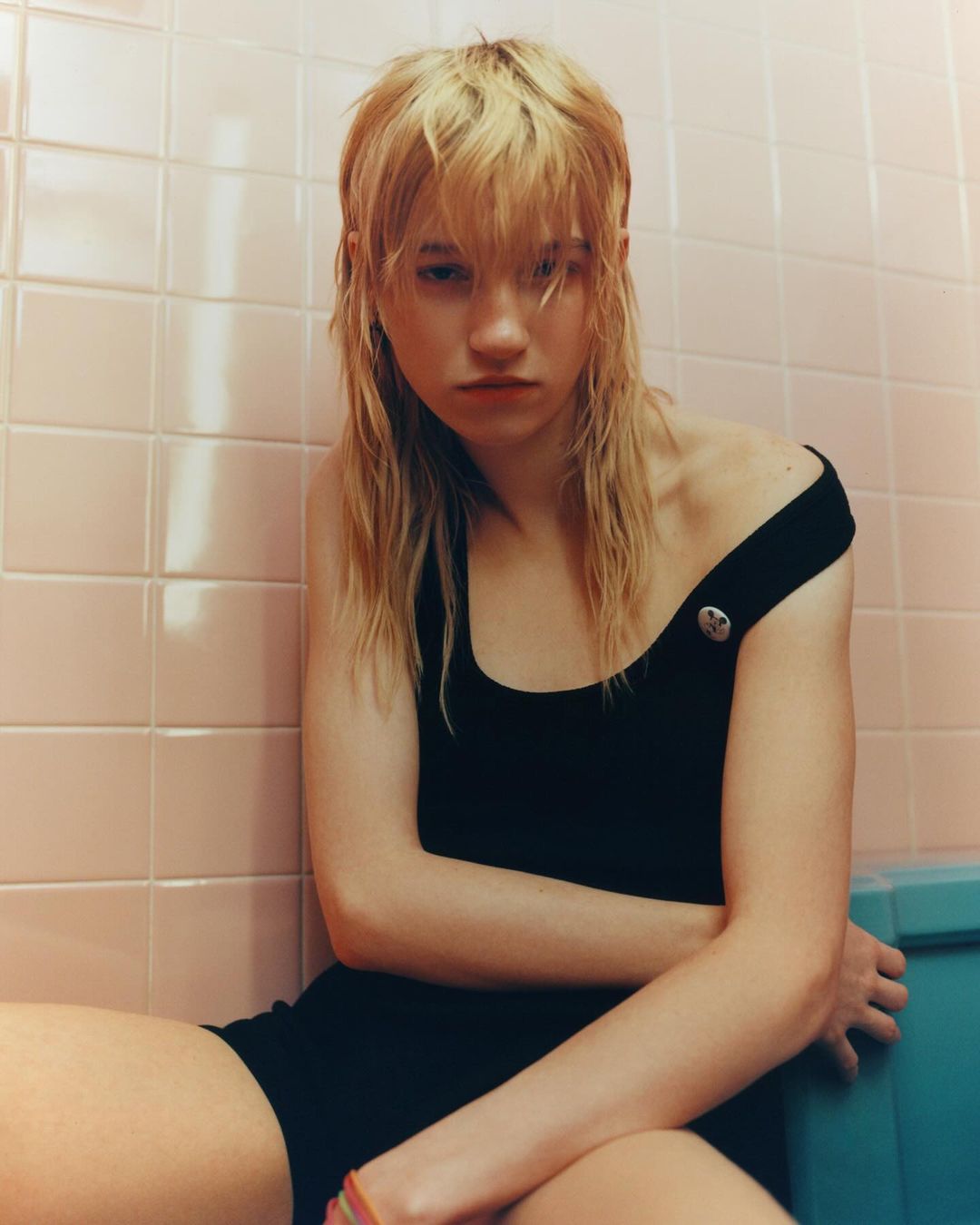 A young adult poses indoors in a black swimsuit against a pink-tiled wall. She has fair skin and blond mullet-style hair.