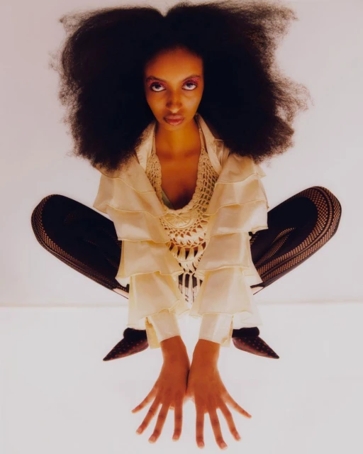 A striking photograph presents an African-American woman with voluminous afro hair, wearing a layered ruffle top, seated on a mirrored chair.