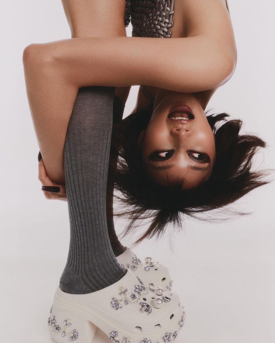 Upside-down portrait of a smiling young woman in gray socks and platform Crocs with embellishments against a white backdrop.