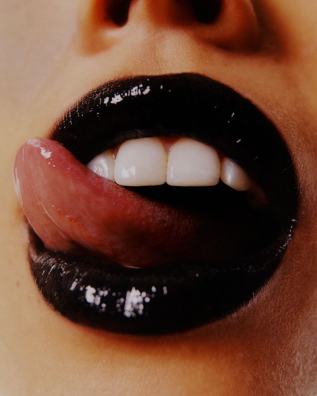 Close-up of a woman's mouth with glossy black lipstick and white teeth, her tongue extended, filling the frame with vivid details.