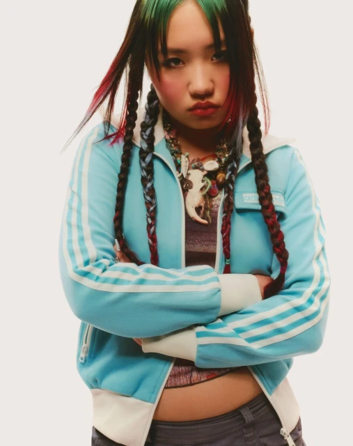 Young woman with multicolored hair, wearing a track jacket and statement necklace, poses with arms crossed against a neutral backdrop.