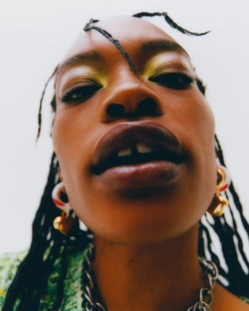 A close-up, low-angle portrait of a woman with dark skin, wearing heavy eye makeup and jewelry, set against a plain white background.