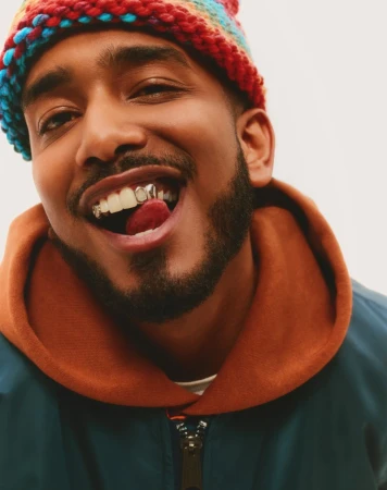 Close-up of smiling adult male with grill, brown hooded sweater and colorful knit hat against a light background in a studio.
