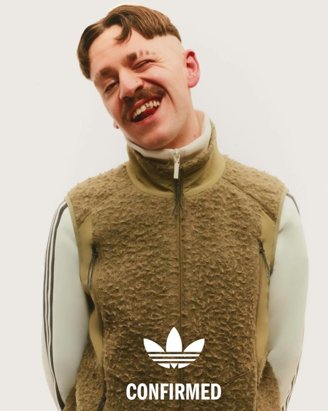A smiling adult male with a mustache and distinct haircut wears an Adidas fleece vest and a white top with Adidas stripes in a studio shot.
