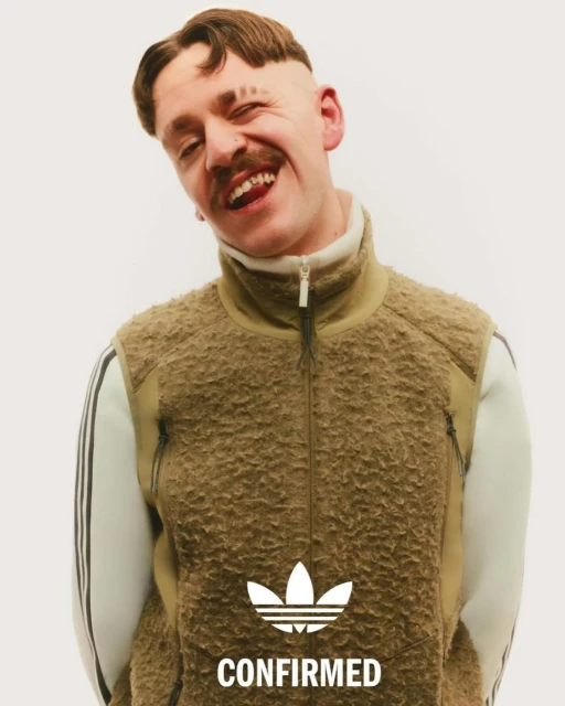 A smiling adult male with a mustache and distinct haircut wears an Adidas fleece vest and a white top with Adidas stripes in a studio shot.