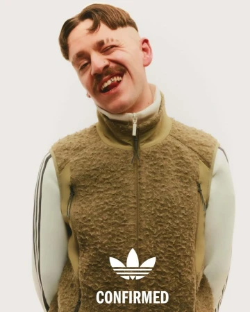 A smiling adult male with a mustache and distinct haircut wears an Adidas fleece vest and a white top with Adidas stripes in a studio shot.