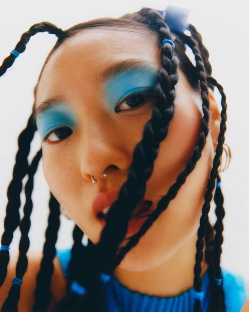 Close-up portrait of a young adult woman with blue eyeshadow and braided hair. The image features a bright, clean aesthetic.