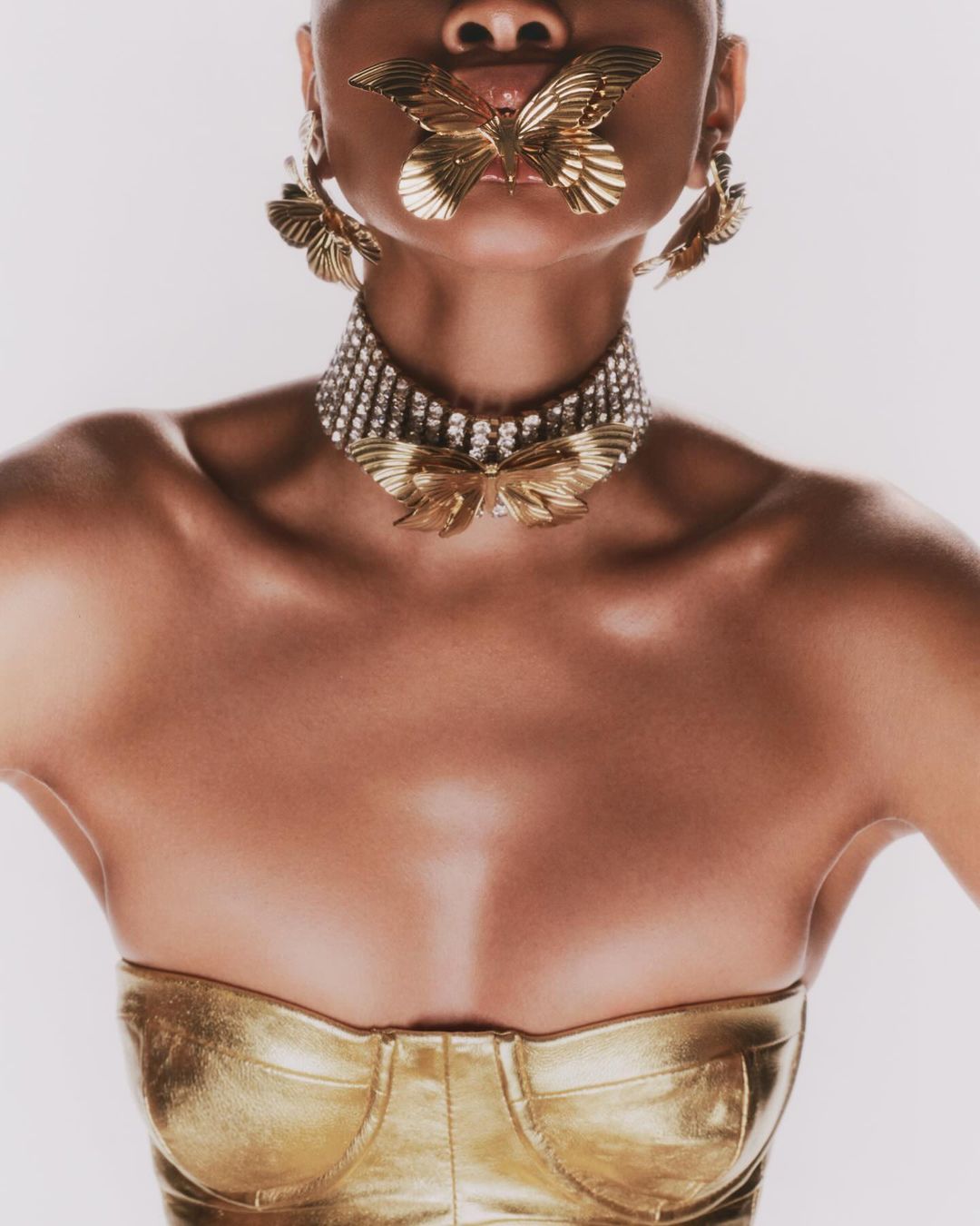 Woman wearing gold bustier and gold butterfly jewelry including earrings, necklace and face accessory against a white backdrop.