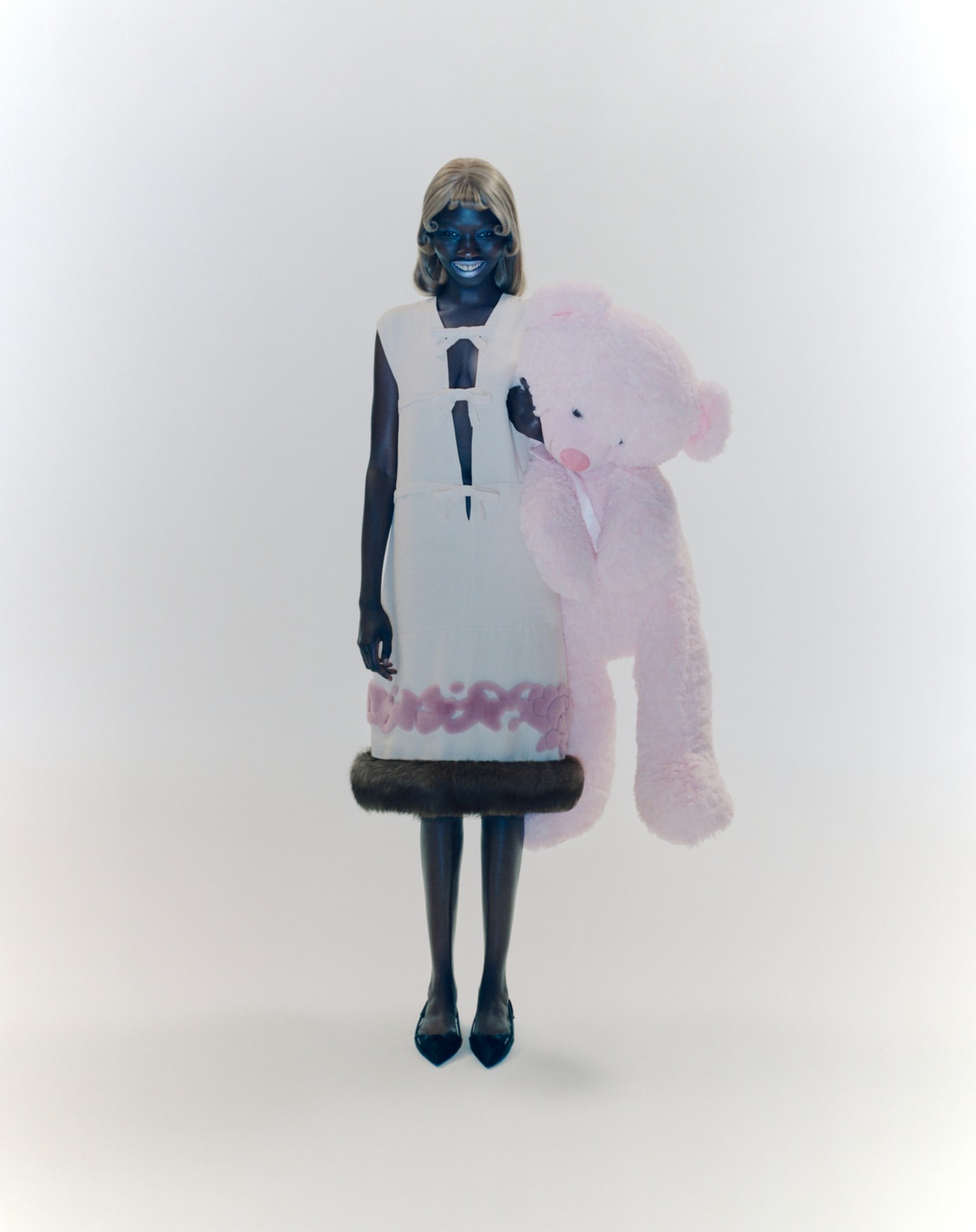 Woman in avant-garde fashion dress and dark shoes holding a large pink teddy bear in a white studio setting, looking at the camera.