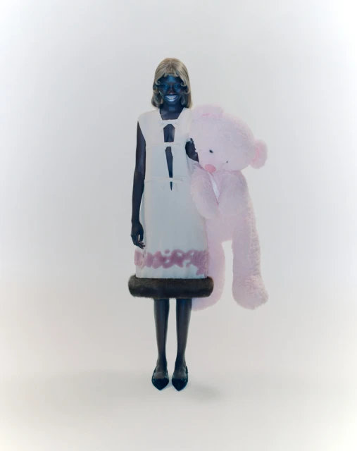Woman in avant-garde fashion dress and dark shoes holding a large pink teddy bear in a white studio setting, looking at the camera.